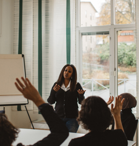 En ung kvinna leder ett möte och sex personer framför henne runt ett bord räcker upp handen.