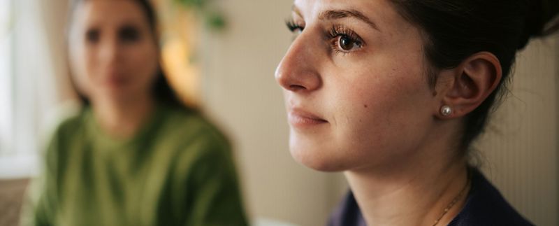Kvinna med sorgsen blick i förgrunden framför en annan kvinna som är suddig.