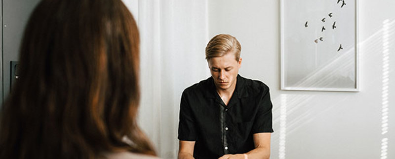 Ung man i samtal med kvinna som bara syns bakifrån. Mannen ser ner på sina händer. Miljön liknar rum för samtalsterapi..