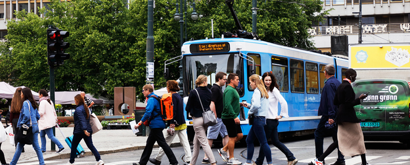 Ett övergångsställe där många människor passerar åt båda håll. I bakgrunden ses en blå buss och en grön skåpbil.