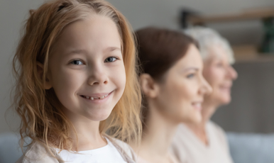 Tre generationer kvinnor, en ung flicka som tittar åt sidan in i kameran, en medelålders och en äldre kvinna som tittar rakt fram. Alla ler. 