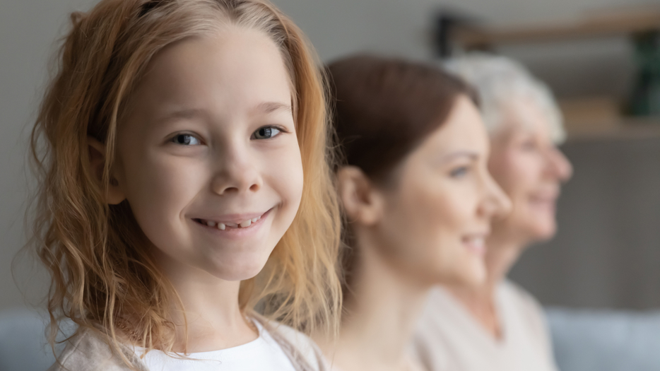 Tre generationer kvinnor, en ung flicka som tittar åt sidan in i kameran, en medelålders och en äldre kvinna som tittar rakt fram. Alla ler. 