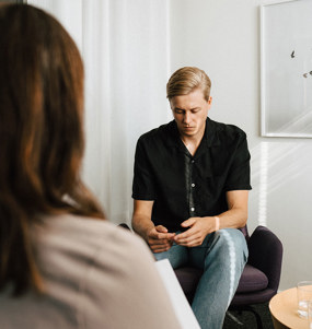 Man med nedböjt huvud sitter mitt emot en psykolog i ett terapirum.