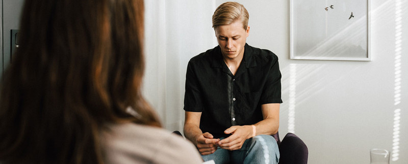 Man med nedböjt huvud sitter mitt emot en psykolog i ett terapirum.