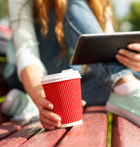Ung kvinna sitter på en parkbänk med en kaffekopp och tittar på en surfplatta. Foto: Mostphotos